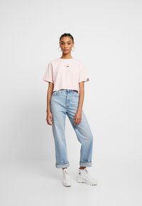 Rozā īsā T-krekls ar priekšējo logotipa dizainu, apvienots ar gaiši ziliem augstās jostasvietas džinsiem un baltiem augstiem sporta apaviem.