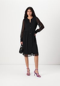 Μαύρο φόρεμα με μακριά διαφανή μανίκια, κουμπιά μπροστά και μαζεμένη μέση. Συνδυασμένο με μωβ σαγιονάρες με λουράκια και μια μικρή μαύρη τσάντα.