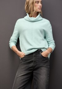 Jasnoniebieski, dzianinowy sweter z kołnierzem typu cowl i ściągaczami, w zestawieniu z ciemnoszarymi, luźno dopasowanymi dżinsami. Modelka stoi z rękami w kieszeniach.