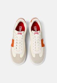 Zapatillas con una parte superior de tela crema con acentos naranjas, detalles en ante beige, cordones blancos y una plantilla roja que muestra el nombre de la marca.
