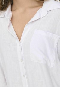 Camicia di lino bianca con colletto, patta di bottoni e taschino sul lato sinistro del petto. La texture è leggera e leggermente sgualcita.
