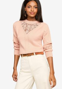 Helles rosa Strickoberteil mit einer Spitze-Einsatz am Halsausschnitt, Zopfmuster-Details an den Ärmeln und gerippten Bündchen. Kombiniert mit weißen Hosen und einem braunen Gürtel.