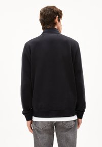 Sweat-shirt en polaire marine avec col et manches longues, présentant un bas côtelé. Porté sur une chemise blanche, associé à un jean gris.