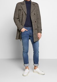 Kappa rutig kappa i beige och brunt, knäppt, bärs över en marinblå polotröja, ihopparad med uppvikta ljusblå jeans och vita sneakers.