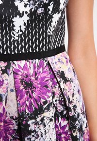 Blumenkleid mit schwarzem und weißem gestreiftem Oberteil, einem plissierten, mehrfarbigen Rock, der mit lila und rosa Blumen verziert ist, und einem schwarzen Gürtel.