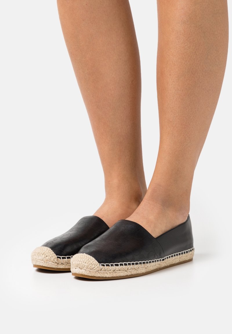 Personne portant des espadrilles en cuir noir avec des semelles en jute tressée beige, debout sur un fond blanc uni.