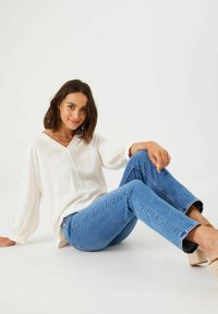 Blusa blanca de corte holgado con mangas largas y escote en V, combinada con jeans de mezclilla azul. El conjunto incluye calzado informal tipo slip-on.