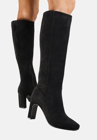 Bottes noires montantes en daim, dotées d'un talon carré et d'un design à bout pointu avec une texture lisse. Détail de zip sur le côté inclus.