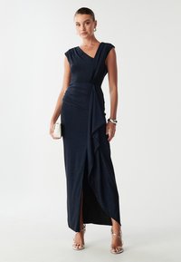 Granatowa sukienka wieczorowa z dekoltem w serek, rękawami cap, marszczonym bodicem i drapowanym przodem. Posiada rozcięcie sięgające ud i gładką teksturę.