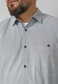 Grijs geruit overhemd met knopen en kraag, voorzien van blauwe knopen en een borstzak met subtiel stiksel. Stoffering met middelfeine structuur.