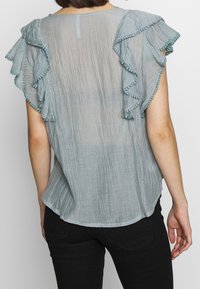 Vaaleansininen, tekstuuroitu paita, jossa on röyhelösateet ja koristeellinen reunus. Hiukan löysä malli, kaareva helma ja pyöreä kaula-aukko.