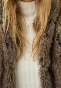 Faux fur scarf in brown tones layered over a ribbed white turtleneck. Scarf features a soft, textured surface with thick fibers.