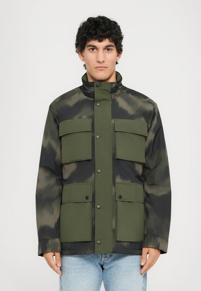 Grüne und schwarze Jacke mit Camouflage-Muster, hohem Kragen und vier großen Taschen auf der Vorderseite; aus glatter Materie gefertigt. Knöpfe entlang der Vorderseite.