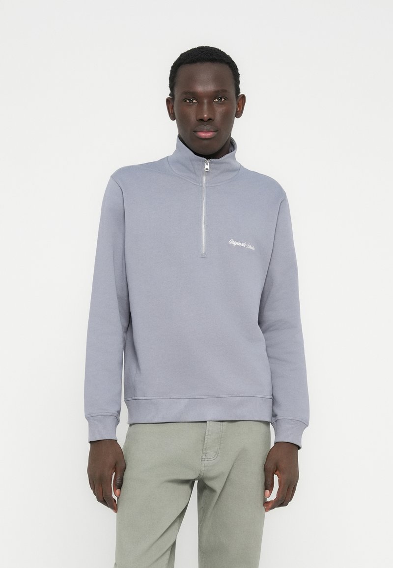 Jasnoszara bluza z kapturem zapinana na zamek błyskawiczny z prążkowanym kołnierzem i mankietami. Na piersi haftowany napis. Noszona z jasnymi zielonymi spodniami.