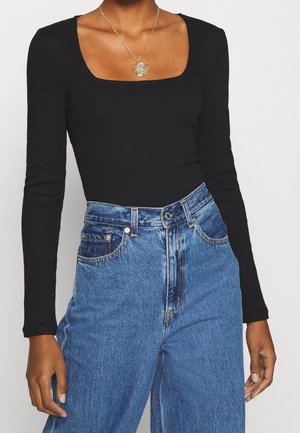 Osoba ubrana w czarną bluzkę z długim rękawem i kwadratowym dekoltem, niebieskie dżinsy z wysokim stanem oraz złoty naszyjnik z zawieszką.