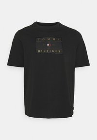 Camiseta de manga corta negra hecha de algodón suave, con un parche de logo rectangular que lleva texto en dorado y blanco, centrado en el pecho.