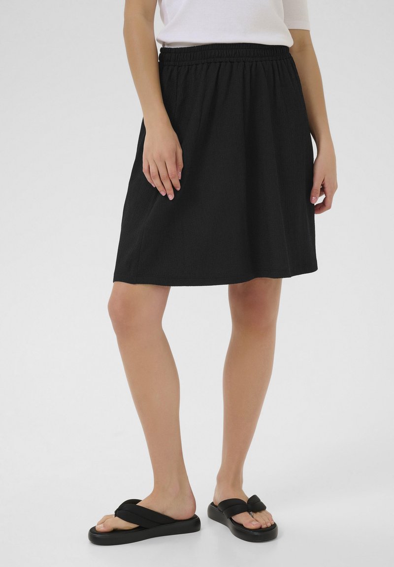 Jupe noire longueur genou avec une taille élastique, présentant un tissu texturé subtil. Associée à des sandales en plastique noires.