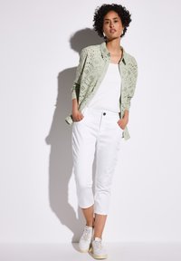 Lichtgroen button-up shirt met een oogjespatroon, gedragen over een witte tanktop. Strakke witte cropped pants. Witte sneakers maken de outfit compleet.