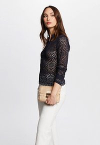 Blusa in pizzo blu con motivo floreale, orlo a scallop e maniche lunghe. Abbinata a una clutch neutra con texture. Pantaloni chiari.