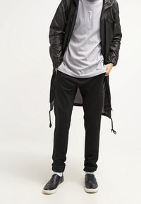 Veste longue noire, légère, avec capuche, portée sur un t-shirt gris avec un motif de chiffres blancs. Pantalon de survêtement noir, baskets foncées et chaussettes blanches.