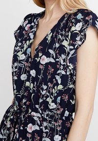 Marinblå blommig klänning med korta volangärmar, prydd med olika blommor i rosa och grönt. Elastisk midja, lätt tyg, v-ringad design.