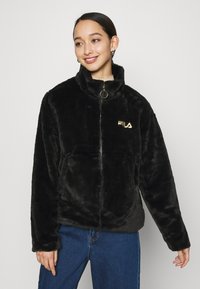 Giacca in finta pelliccia nera con colletto alto, chiusura a zip e logo dorato. Presenta una texture morbida e una vestibilità comoda, abbinata a jeans blu.