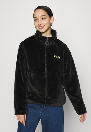 Giacca in finta pelliccia nera con colletto alto, chiusura a zip e logo dorato. Presenta una texture morbida e una vestibilità comoda, abbinata a jeans blu.