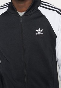 Černobílá zipová bunda Adidas s vysokým límcem, s třemi bílými pruhy na ramenou a vyšitým logem na hrudi.
