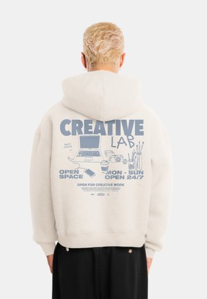 Person med kort blondt hår iført en hvid hættetrøje med blå "Creative Lab" tekst og illustrationer af bærbar, kaffe og kunstmaterialer.