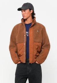 Brown fleece-lined jacket with a quilted texture and snap closure, paired with a navy t-shirt and black jeans. Includes a grey cap.
