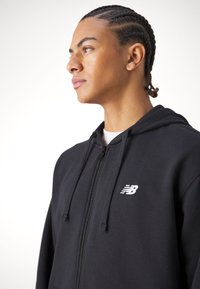 Melna kokvilnas kapuce, kas aizdares ar rāvējslēdzēju, ar priekšējo kabatu un baltu New Balance logo uz krūšu zonas. Gluda tekstūra, regulējama kapuces aukla.