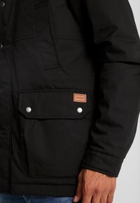 Chaqueta negra con una textura suave, que cuenta con un gran bolsillo frontal, dos cierres a presión y un parche con logo. Diseñada con un cuello alto y costuras resistentes.