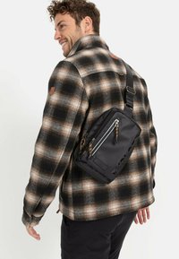 Zwarte schoudertas met twee met rits afsluitbare vakken, een textuur met uitsparingen en een verstelbare band, gedragen over een geblokt flanellen shirt.