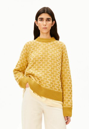Jonge vrouw met lang donker haar, die een gele trui met patroon en licht beige broek draagt, staand met één hand in de zak tegen een witte achtergrond.