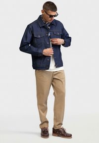 Chaqueta de mezclilla en azul profundo con botones en el frente, dos bolsillos en el pecho y costuras en naranja contrastante. Combinada con pantalones beige y zapatos marrones.