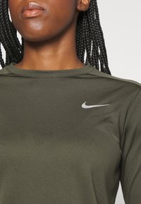 Nahaufnahme einer Person, die ein olivgrünes Nike-Shirt mit einem kleinen weißen Swoosh-Logo auf der oberen Brust trägt und geflochtene Haare sichtbar sind.