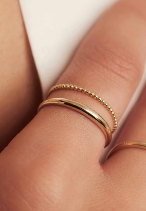 Trois bagues en or sur une main : une lisse, une avec un design en boule arrondie, et une anneau simple. Le teint de la peau est chaud.