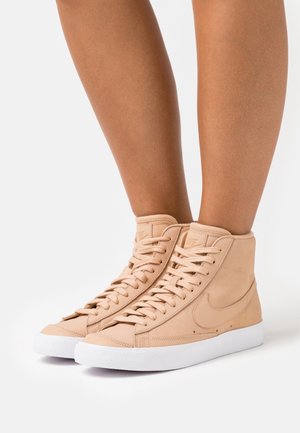 High-top sneakers in nude leather with a textured finish, featuring white rubber soles, laces, and a subtle logo on the side.