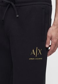 Μαύρη φόρμα με πλαϊνή τσέπη και χρυσλό κεντημένο λογότυπο "A|X Armani Exchange" κοντά στον μηρό, φορεμένη από άτομο με εμφανές χέρι.