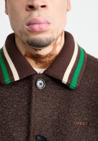 Brun tekstureret jakke med ribbet krave med grønne og cremefarvede striber, sort knaplukning og "OBEY" logo detaljer.