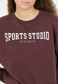 Bordeaux Sweatshirt aus weichem Stoff mit weitem Halsausschnitt, versehen mit dem auffälligen weißen Text "SPORTS STUDIO" und "ATHLECIA" in der Mitte.