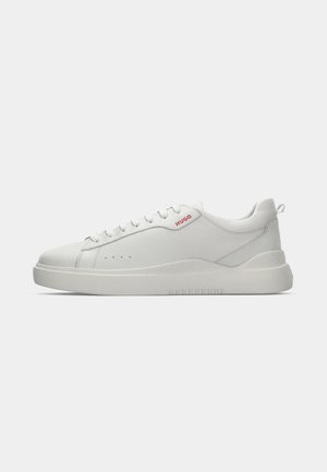 White sneaker with a smooth leather finish, round toe, four eyelets, subtle perforations, and a red logo detail on the side.