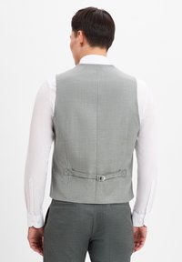 Graham textured Weste von hinten betrachtet, mit einem verstellbaren Schnallenband und einer glatten Oberfläche, kombiniert mit einem weißen Langarmshirt.
