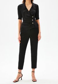 Blouse noire à manches courtes, au design ajusté, avec taille froncée et trois boutons décoratifs. Associée à un pantalon noir ajusté et des talons à lanières.