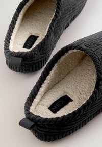 Black fabric slippers with plush beige lining, ribbed texture on the exterior, and a small fabric loop at the back for easy wear.