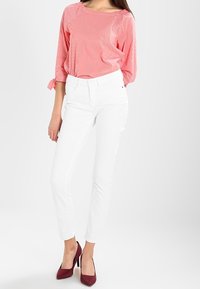 Blouse rayée corail à manches trois-quarts, associée à un pantalon blanc ajusté et des talons bordeaux. La blouse a un col rond et se noue aux manches.