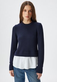 Pull en tricot bleu marine avec des manches longues et un col côtelé, superposé sur une chemise blanche à boutons qui dépasse en dessous, associé à un jean bleu.