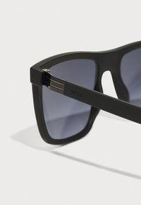 Ochelari de soare rectangulari negri cu lentile gri închis. Dispune de rame texturate mate și detalii argintii pe balamale. Logo pe lateral.