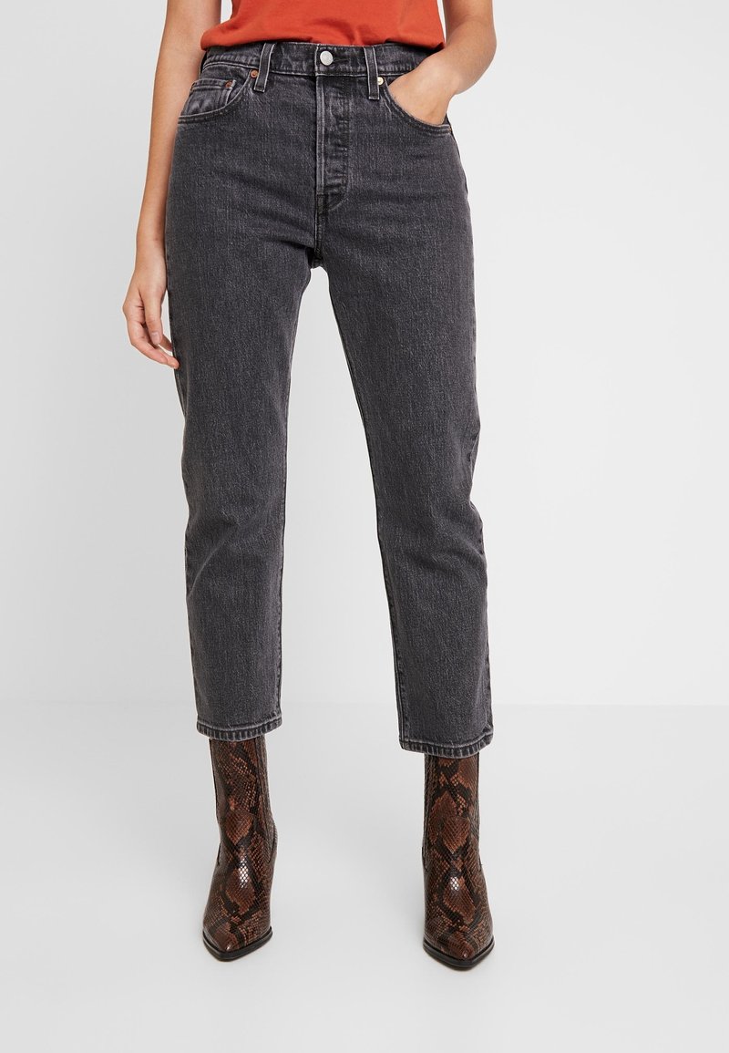 Personne portant un jean en denim noir taille haute et des bottes à bout pointu à motif peau de serpent marron, avec une main dans la poche.