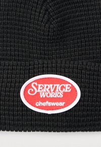 Μαύρο ριμπ σκουφί με κόκκινο και λευκό οβάλ έμβλημα με το κείμενο "SERVICE WORKS chefswear." Απαλή υφή, εφαρμοστή.